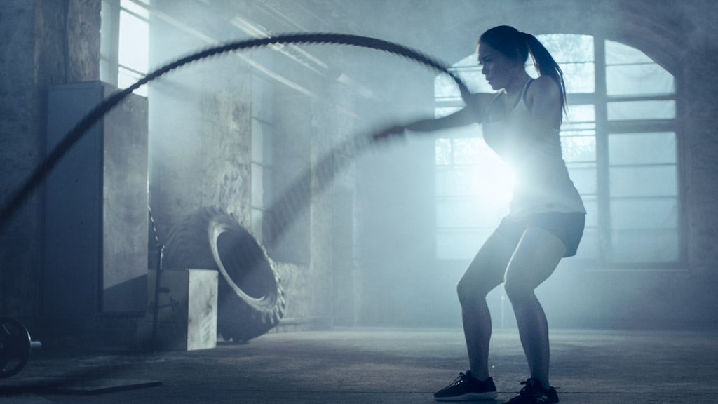 Strong Athletic Woman Exercises with Battle Ropes as Part of Her Cross Fitness Gym Workout Routine. She's Covered in Sweat and Training Takes Place in a Abandoned Factory Remodeled into Gym.